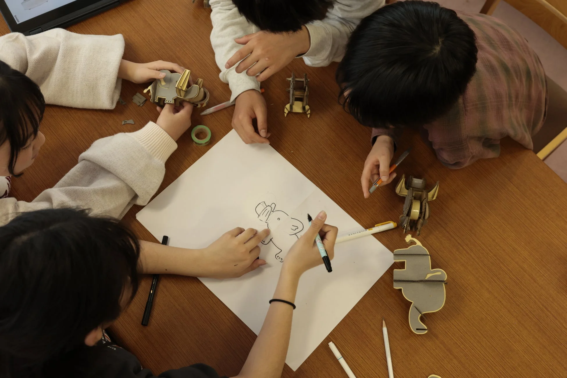 子供たちがテーブルを囲み、紙に絵を描いたり、段ボール製の動物の模型を組み立てたりして、創造的な活動を楽しんでいる様子