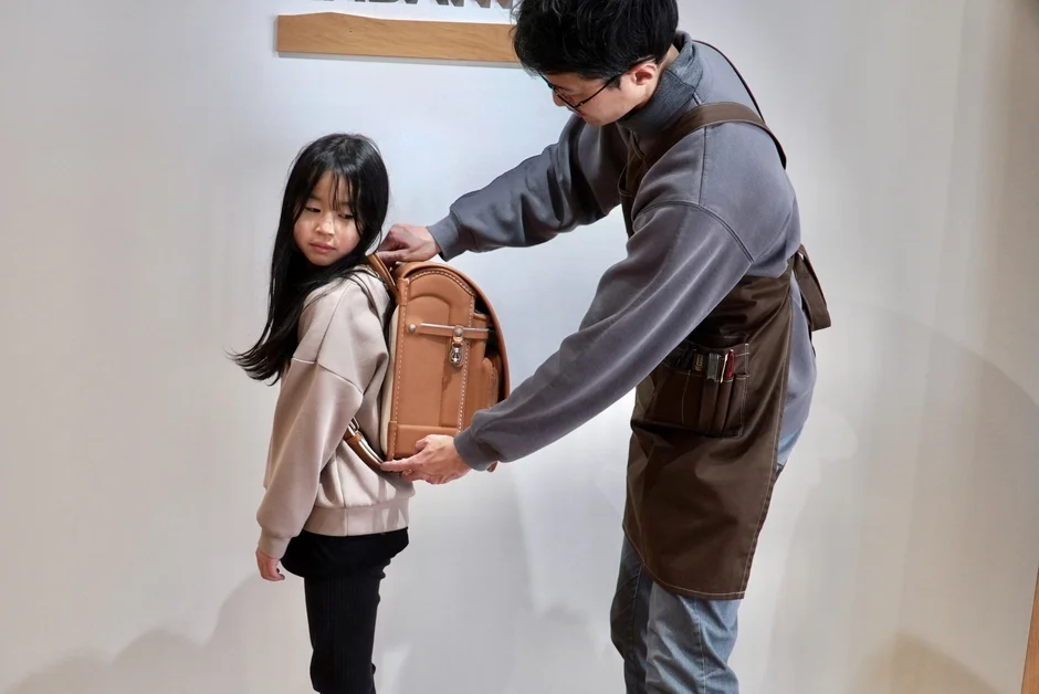 エプロンをつけた男性が、小さな女の子に茶色のランドセルを背負わせてフィッティングしている様子