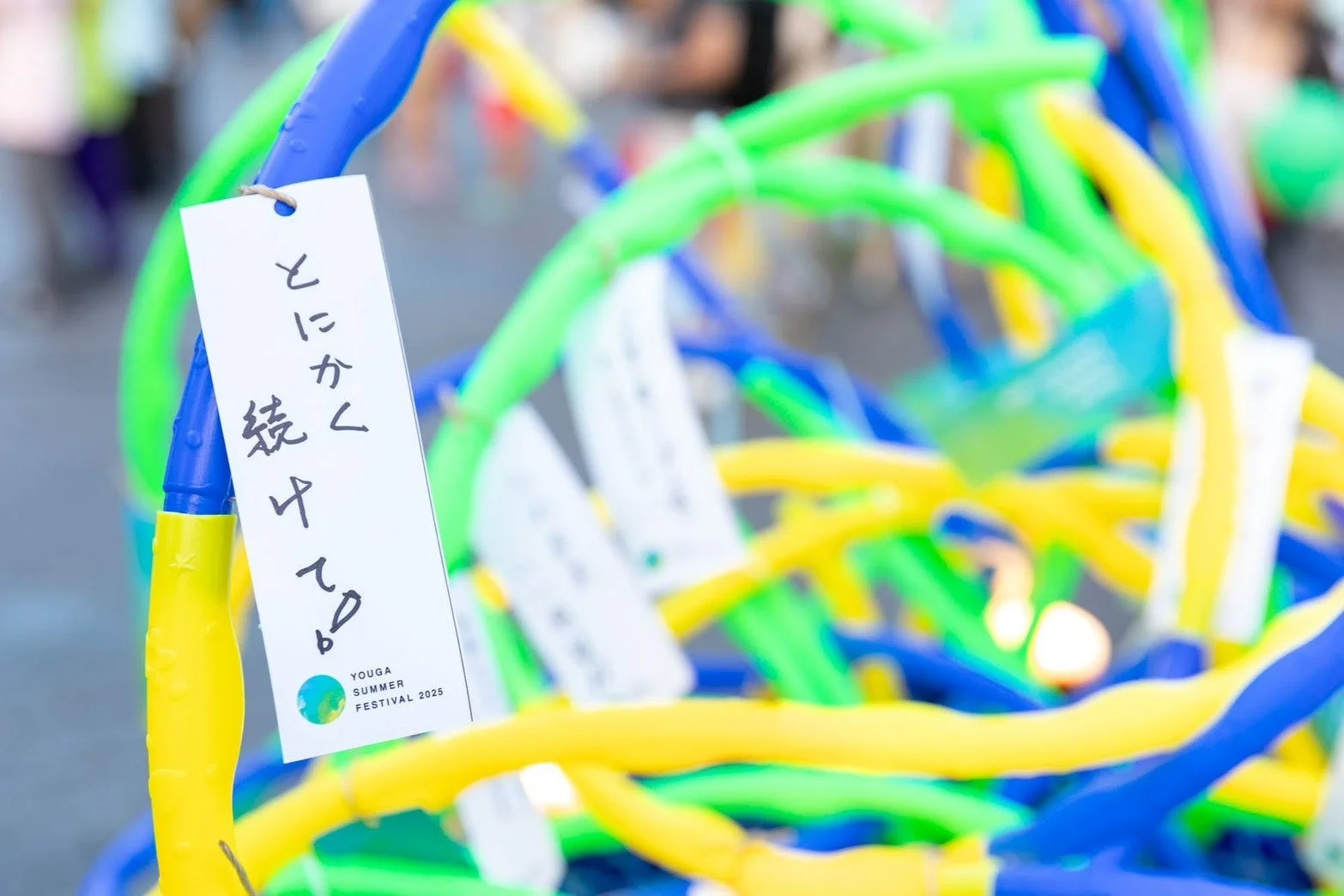 「とにかく続けてる!」と書かれた短冊が、青、黄、緑のカラフルなチューブ状の飾り付けに吊るされている夏のイベント風景。