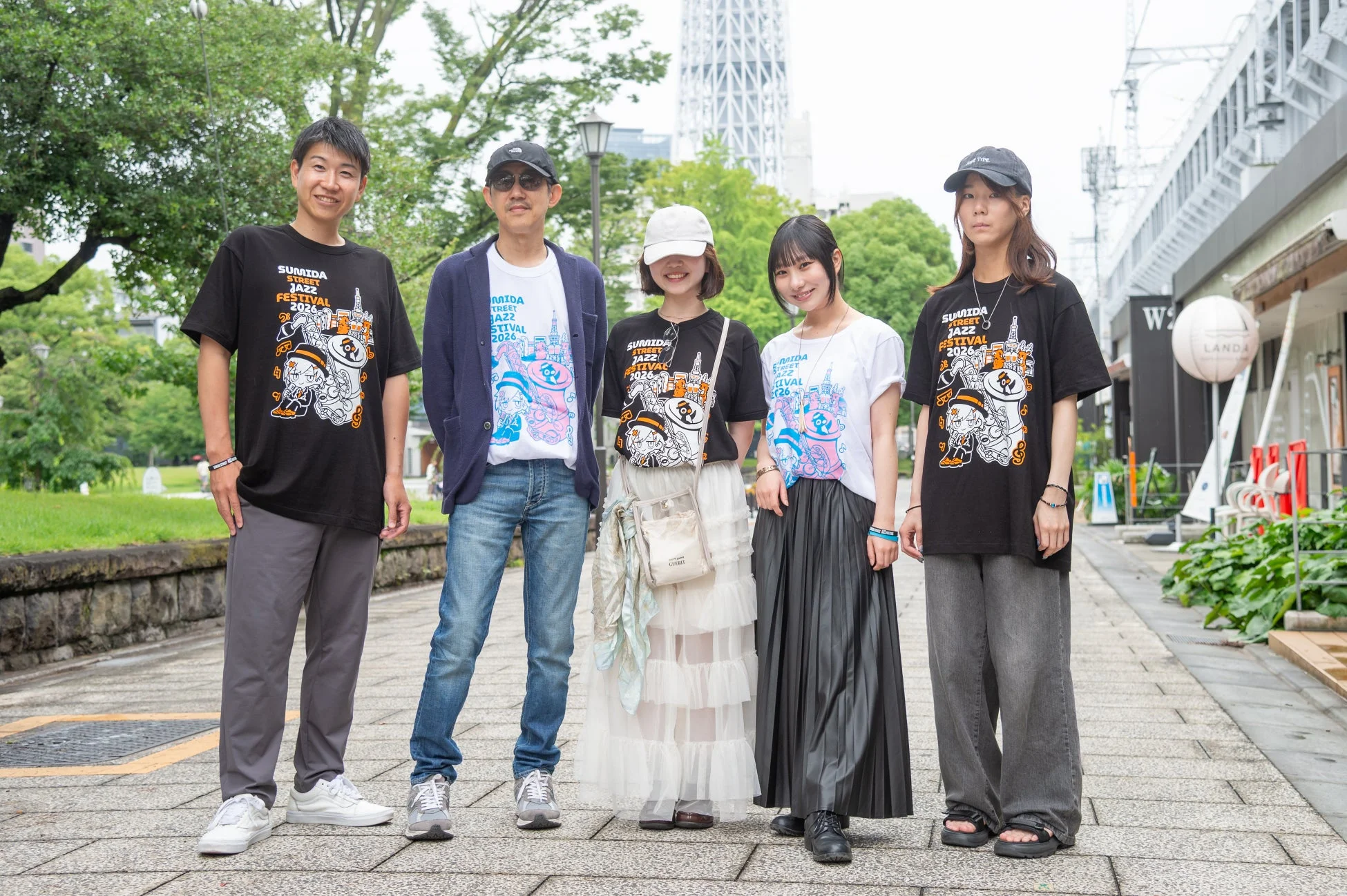 墨田ストリートジャズフェスティバルのTシャツを着た男女5人が屋外で笑顔で立っている写真。背景には木々や建物、東京スカイツリーが見える。フェスティバルの関係者か参加者と思われる。