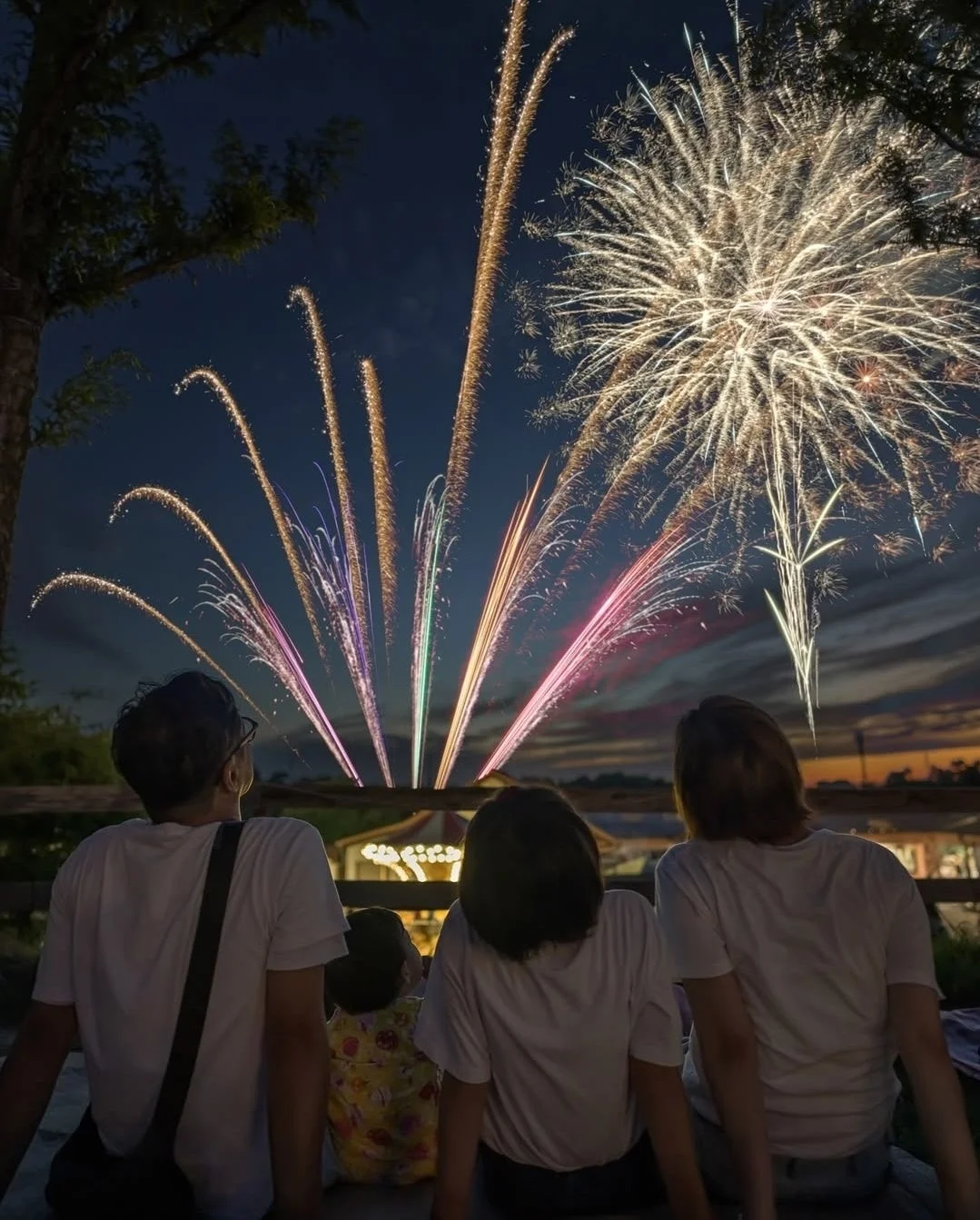 夕暮れの空に打ち上がる色とりどりの花火を、家族が後ろ姿で鑑賞している様子です。夏の夜の思い出を彩る美しい光景が広がっています。