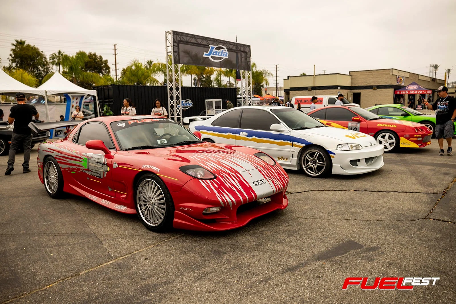 屋外のイベント会場で、赤色のマツダRX-7をはじめとするカスタマイズされたスポーツカーが複数展示されています。人々が集まり、様々なブランドロゴやイベント名「FUEL FEST」が見えます。