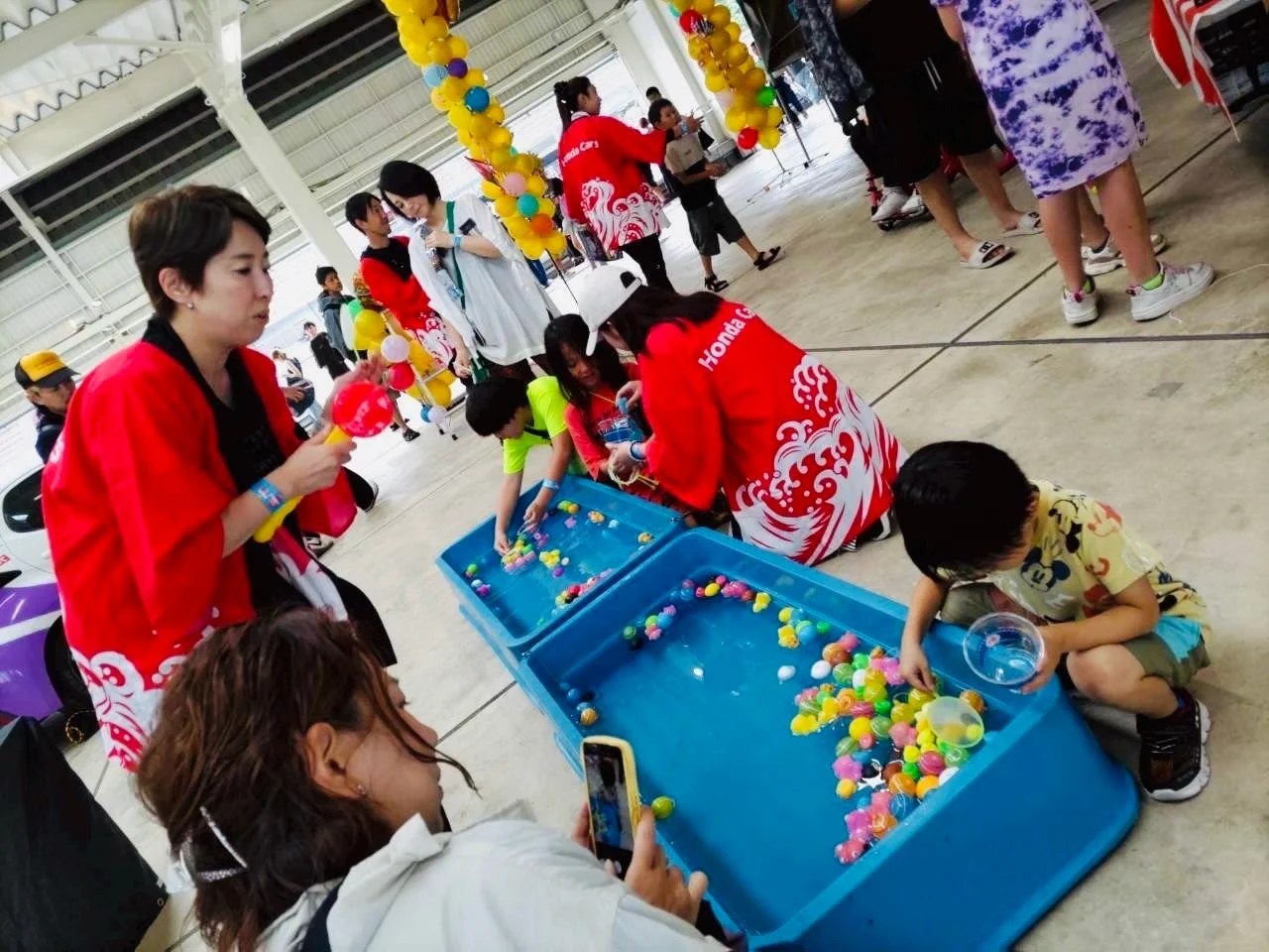 屋内で行われているイベントで、子供たちが青い容器に浮かぶカラフルなボールや魚のおもちゃをすくって遊んでいます。