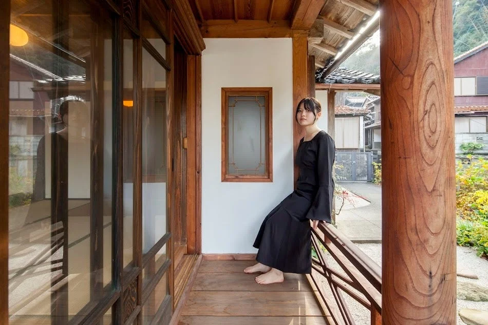 黒いドレスを着た若い女性が、日本の伝統的な木造家屋の縁側に座っているポートレート