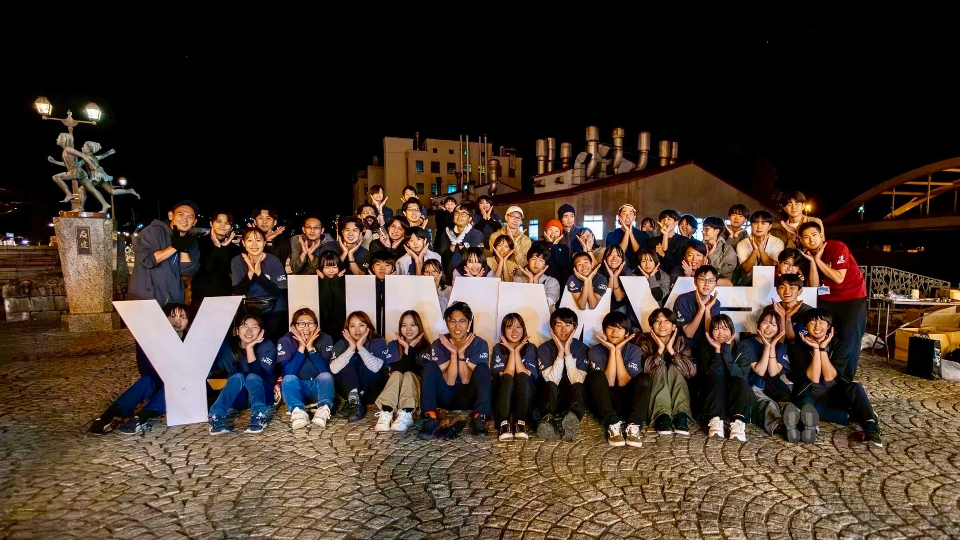 夜間、大勢の日本人が「YUMESAKI」と書かれた大きな白い文字の周りに集まり、笑顔でポーズをとっている集合写真です。多くの人が頬に手を当てるポーズをしており、イベントか何かの集まりの様子がうかがえます。背景には都市の建物や橋が見えます。