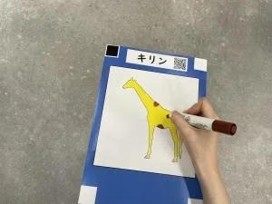 青いボードに貼られたキリンの絵に、茶色のマーカーペンで色を塗っている手の様子を捉えた画像です。キリンは黄色く塗られ、斑点が描かれており、教育的な活動やクリエイティブな遊びの瞬間を示唆しています。