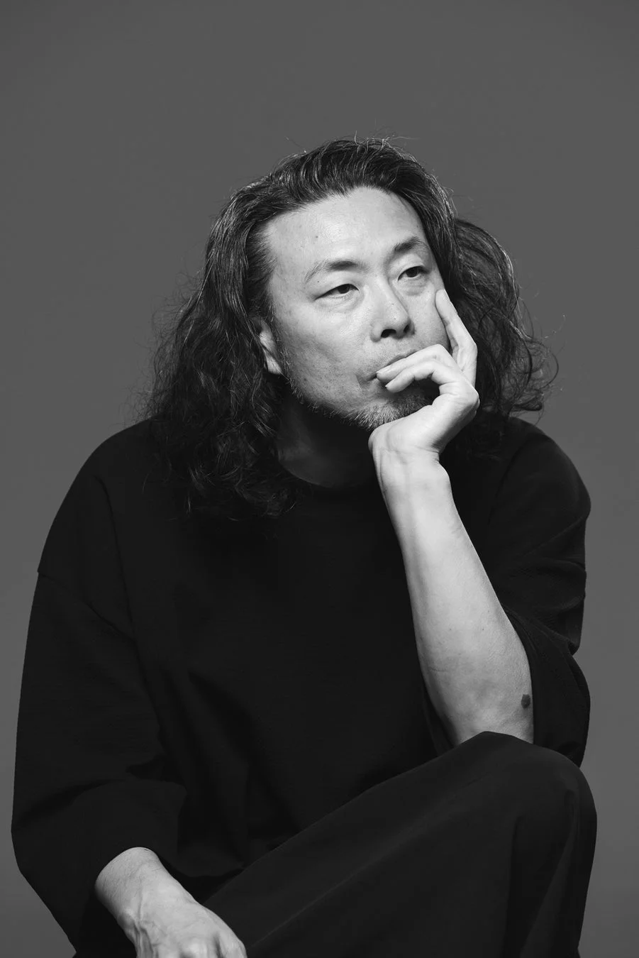 長髪と髭を生やしたアジア系の男性が、顔に手を当てて思案している様子のモノクロポートレート