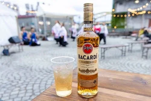 木製のテーブルにバカルディ・カルタ・オロのラム酒のボトルと飲み物が入ったプラスチックカップが置かれている。背景には屋外イベントらしき場所で人々が写っている。