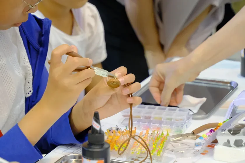 子供がレジン液や工具を使ってアクセサリー作りに集中している様子。テーブルには様々なビーズや材料が並び、手作りの楽しさが伝わるワークショップ風景です。