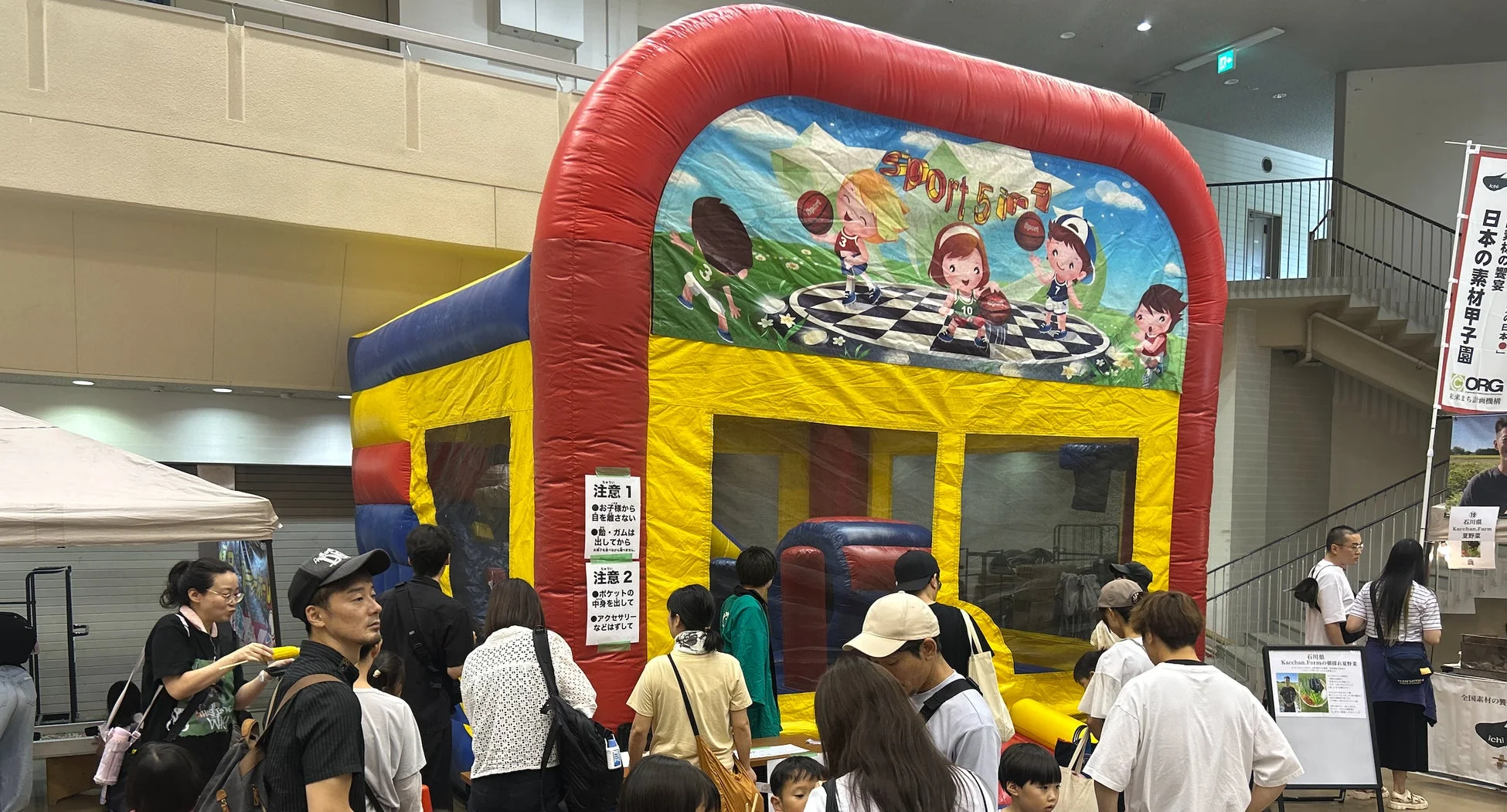 屋内イベント会場で、スポーツテーマの大型インフレータブル遊具が設置され、多くの家族連れで賑わっている。