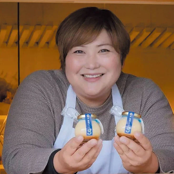 エプロンを着た女性が、満面の笑みで美味しそうな瓶入りプリンを両手に持っています。彼女の陽気な表情が、そのデザートの魅力を一層引き立てています。