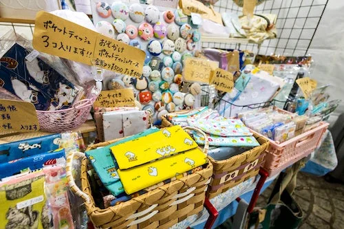 クラフトマーケットの屋台で、手作りの布小物が豊富に並んでいます。猫や鳥柄のポケットティッシュケースやポーチが中心で、賑やかな販売風景がうかがえます。