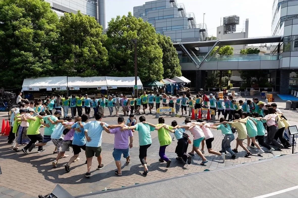 晴れた屋外の広場で、多くの人々が肩を組み、輪になって踊るかのような共同作業を楽しんでいます。