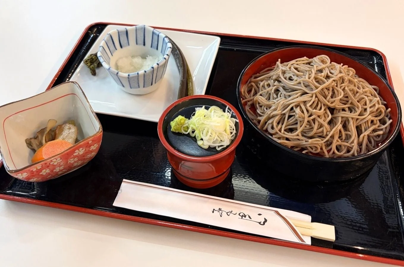 黒と赤のお盆に盛り付けられた和食の定食です。メインは蕎麦で、薬味としてわさびと刻みネギ、大根おろし、そして煮物などの小鉢が添えられています。