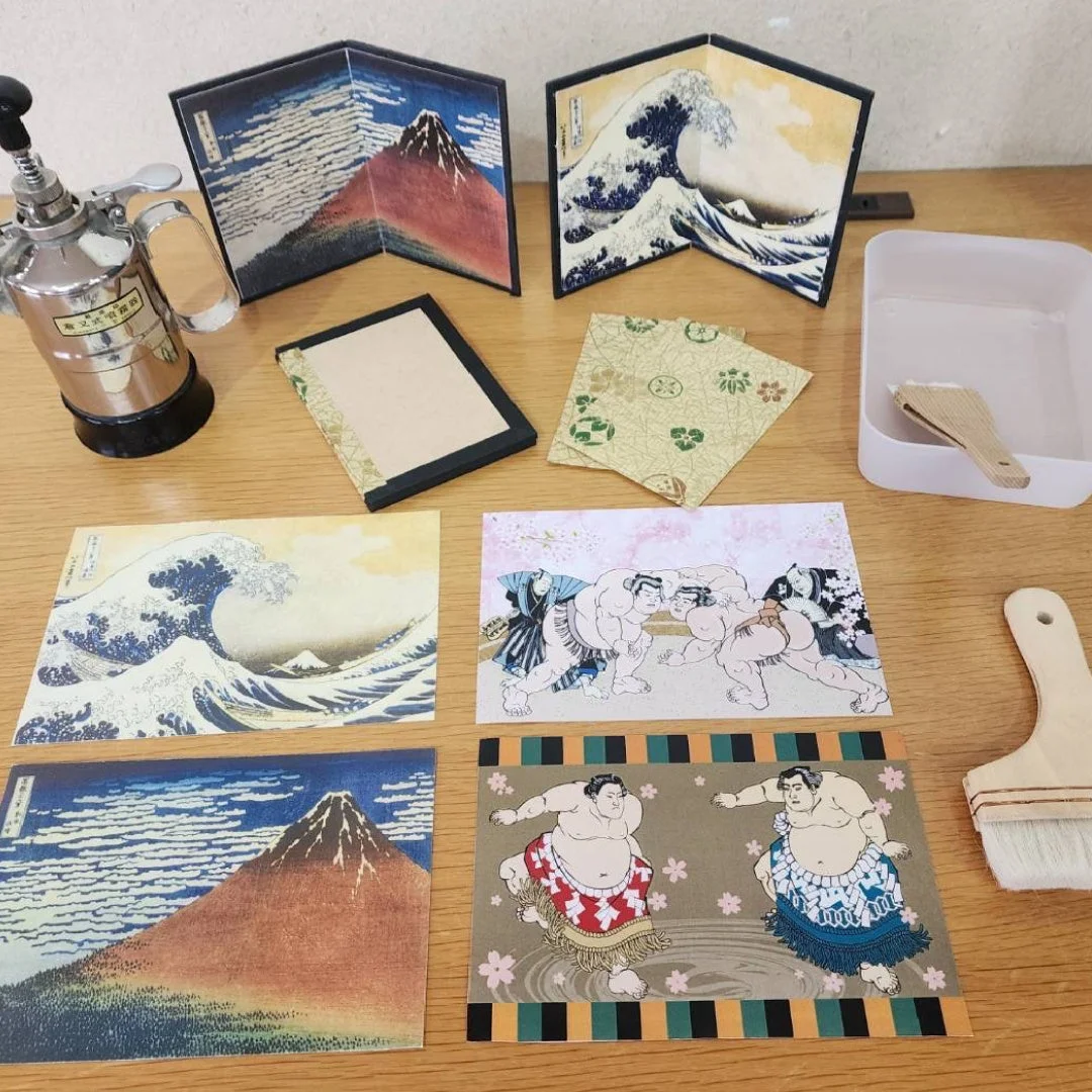 葛飾北斎の浮世絵(富嶽三十六景の神奈川沖浪裏、凱風快晴)や相撲力士が描かれたポストカードやミニ屏風、そして文明開化と記されたスプレー缶や筆などのクラフト用品が木製のテーブルに並べられています。