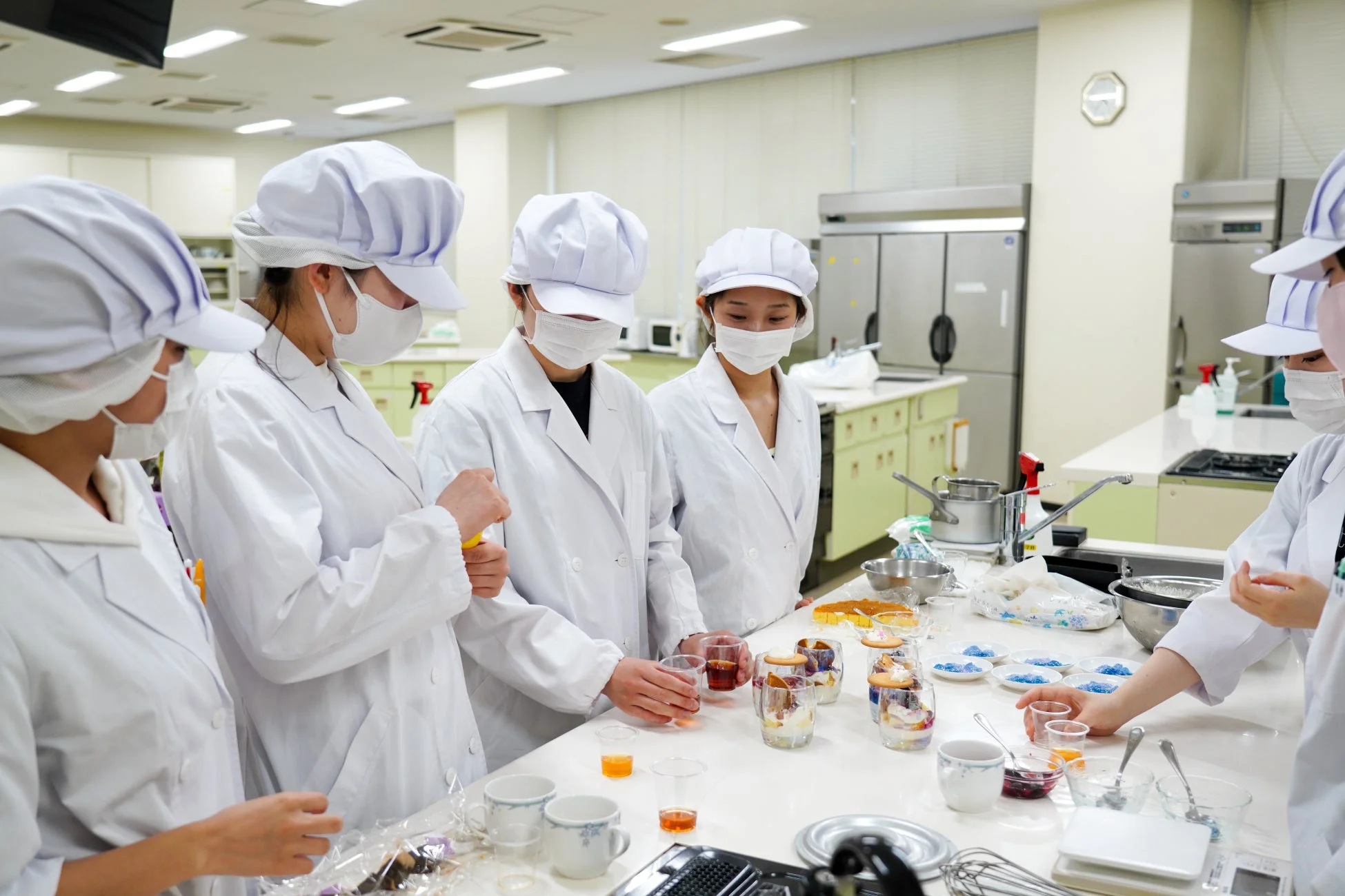 白衣とマスクを着用した学生たちが厨房で調理実習や試食を行っている様子