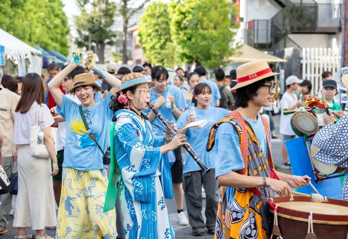 夏祭りのパレードで、浴衣を着た女性がクラリネットを演奏し、他の参加者も太鼓を叩きながら笑顔で楽しんでいる。青いTシャツを着た多くの若者が集い、活気あるお祭りの雰囲気が伝わる一枚。