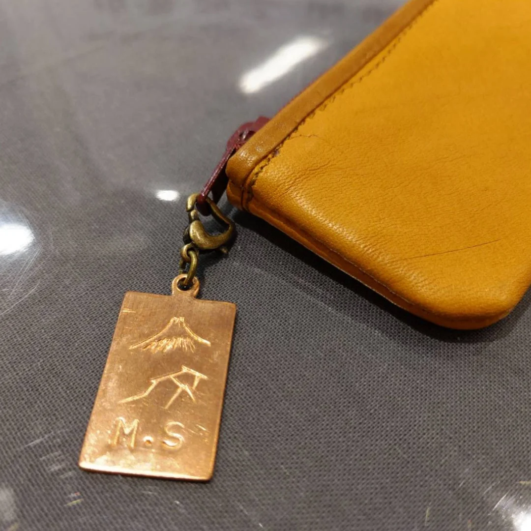 黄色の革製ポーチに、富士山とイニシャル「M.S」が刻印された銅色の金属製チャームが取り付けられています。
