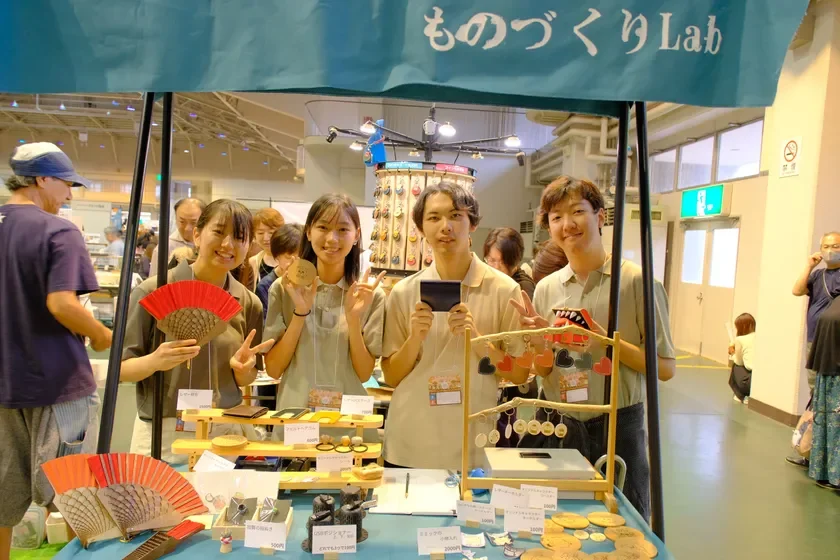 ものづくりLabのブースで、若い男女4人が笑顔で手作りの扇子や革製品などを紹介しています。様々なクラフト品が並び、賑やかなイベントの様子がうかがえます。