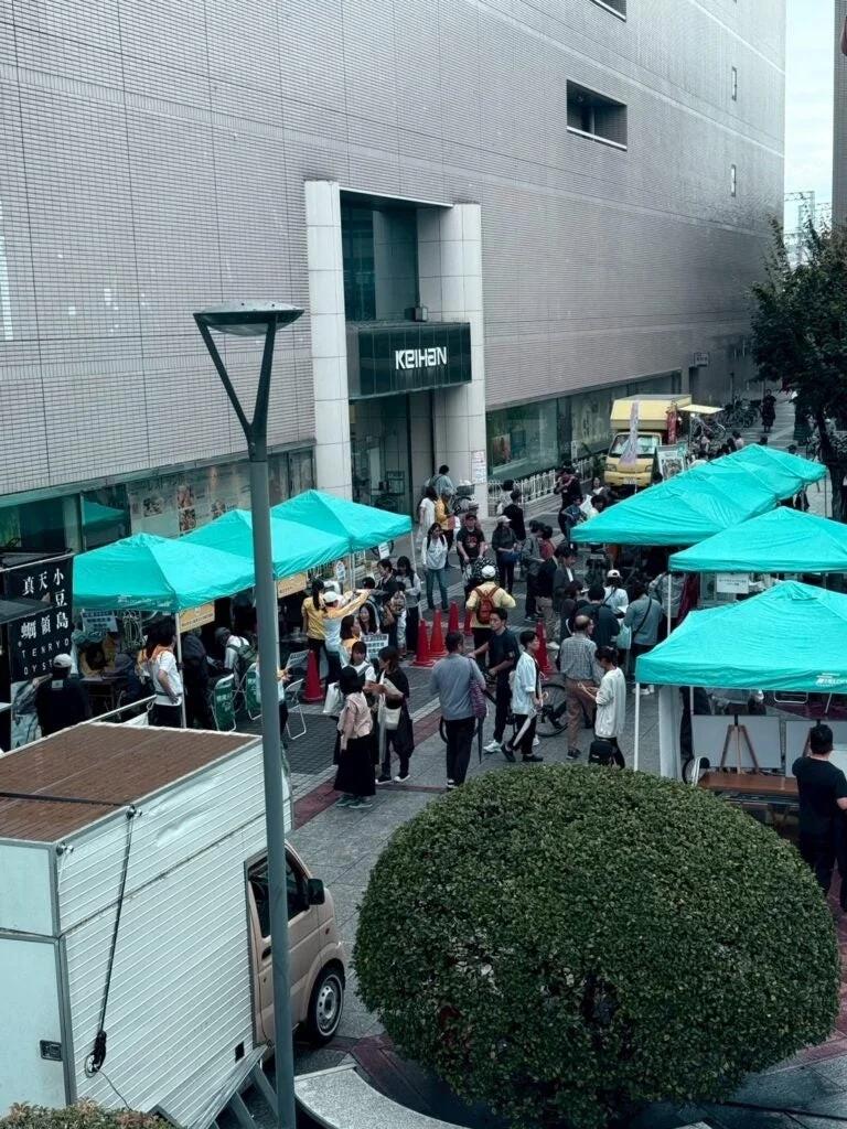 京阪の建物前で賑やかなストリートマーケットが開催されている様子。多くの人々が緑色のテントが並ぶ屋台を訪れ、活気あふれるイベントを楽しんでいる。