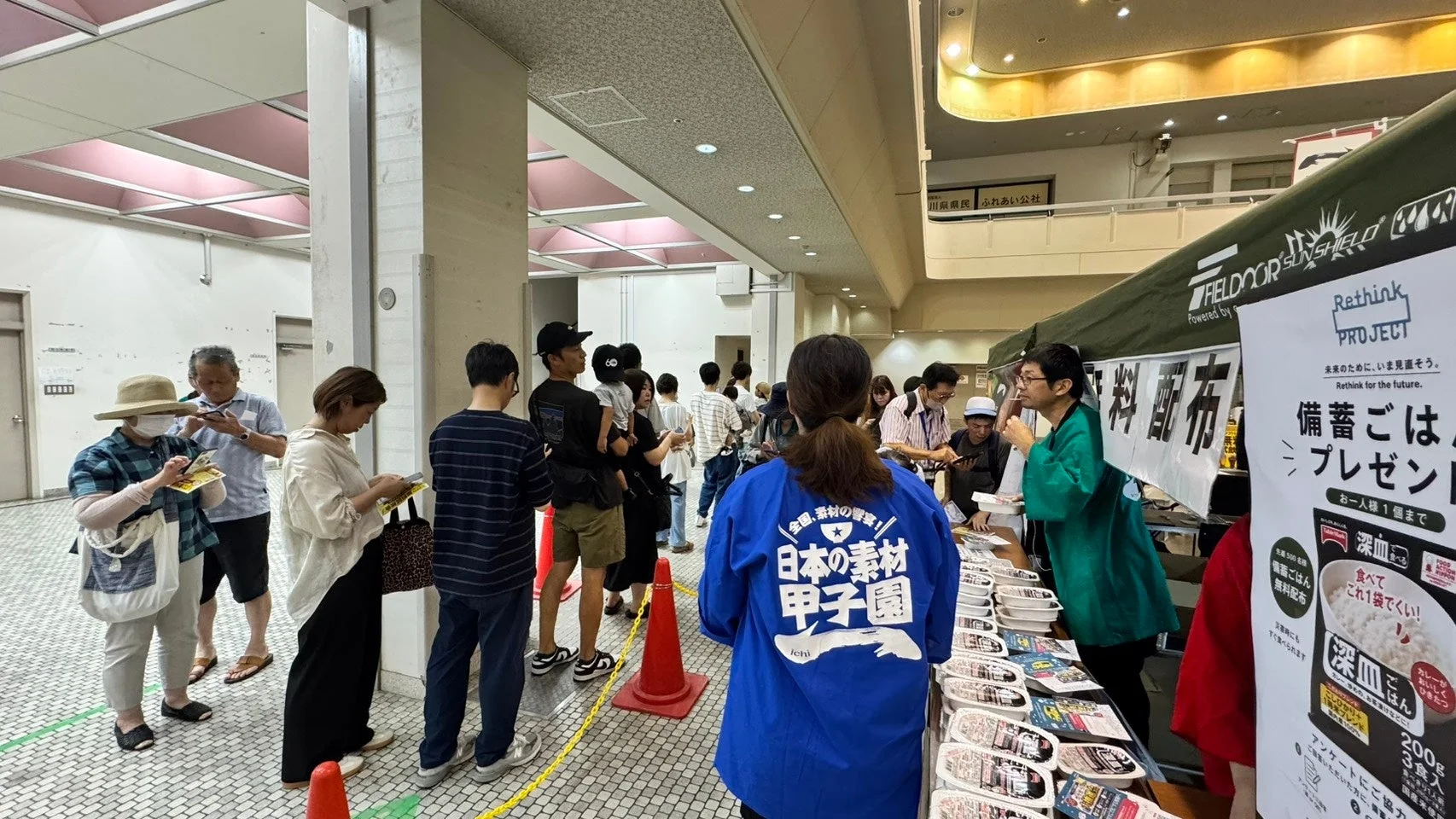 屋内で開催されているイベントで、多くの人々が「Rethink PROJECT」による備蓄ごはんの無料配布に並んでいます