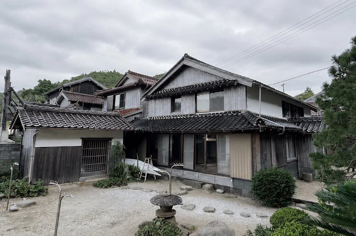 瓦屋根と木造の伝統的な日本家屋が立ち並び、手前には石灯籠のある砂利の庭が広がる風景