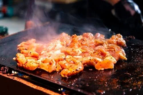 熱い鉄板の上で、香ばしい煙を上げながら焼かれている鶏肉と思われる肉片のクローズアップ。食欲をそそる調理風景が捉えられています。