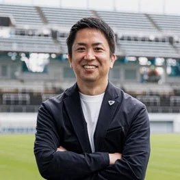 スタジアムを背景に、ネイビーのジャケットを着た男性が腕を組み、カメラに向かって笑顔を見せています。
