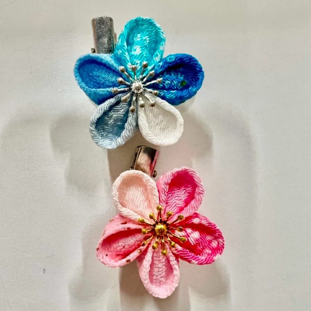 白い背景に、青系とピンク系の花をモチーフにしたつまみ細工のヘアクリップが2つ並べられています。
