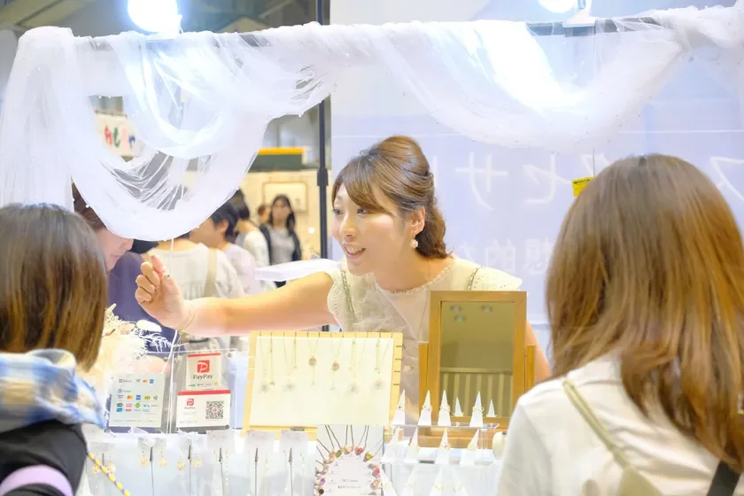イベント会場のブースで、笑顔の女性店員がお客様にハンドメイドアクセサリーを説明・販売している様子。ネックレスやイヤリングが並び、PayPayなどの支払い方法が示されている。