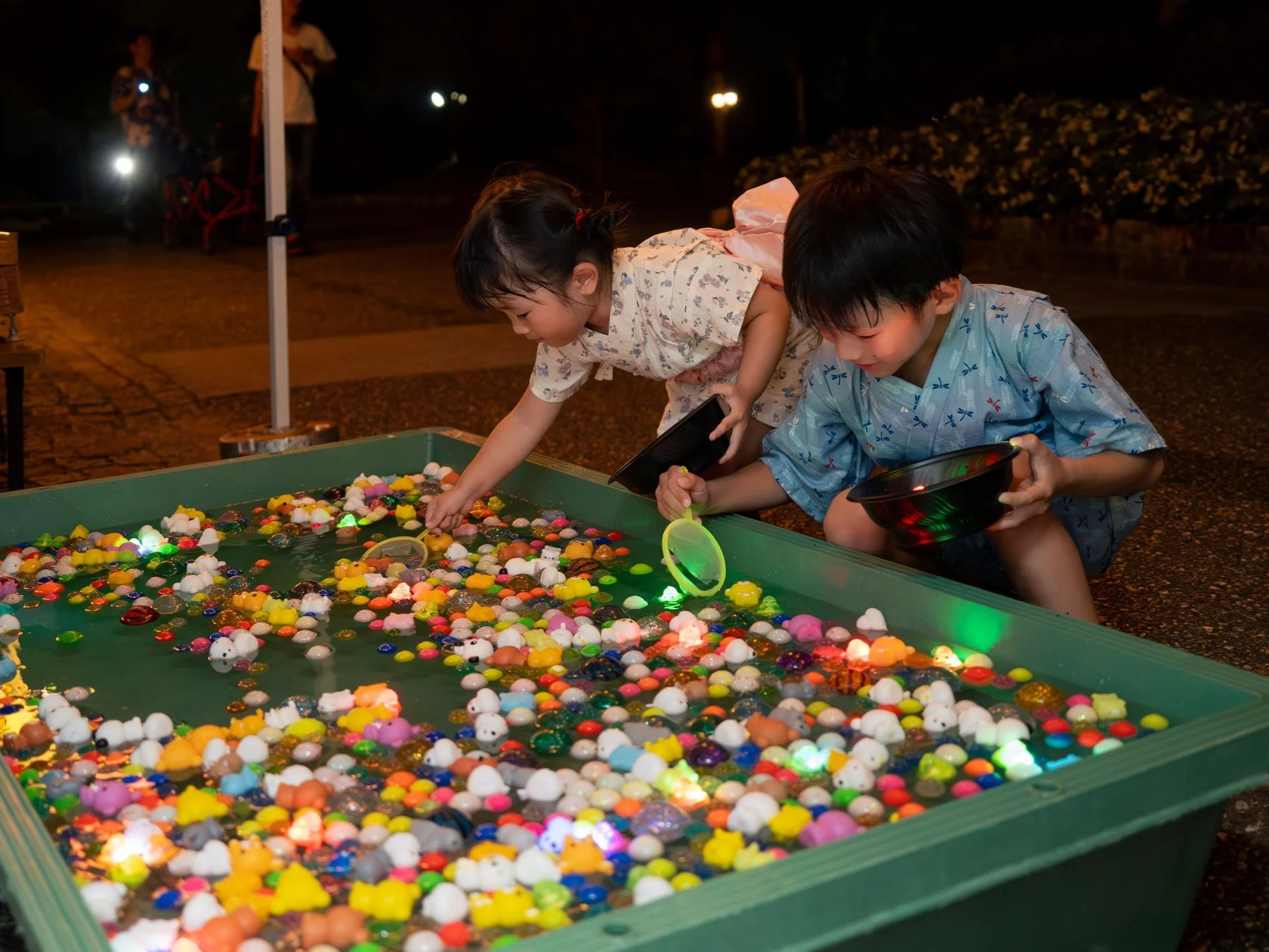 夜の夏祭りで、浴衣を着た二人の子供が光るおもちゃやカラフルなゴム製のおもちゃが浮かぶ水槽でおもちゃすくいを楽しんでいます。