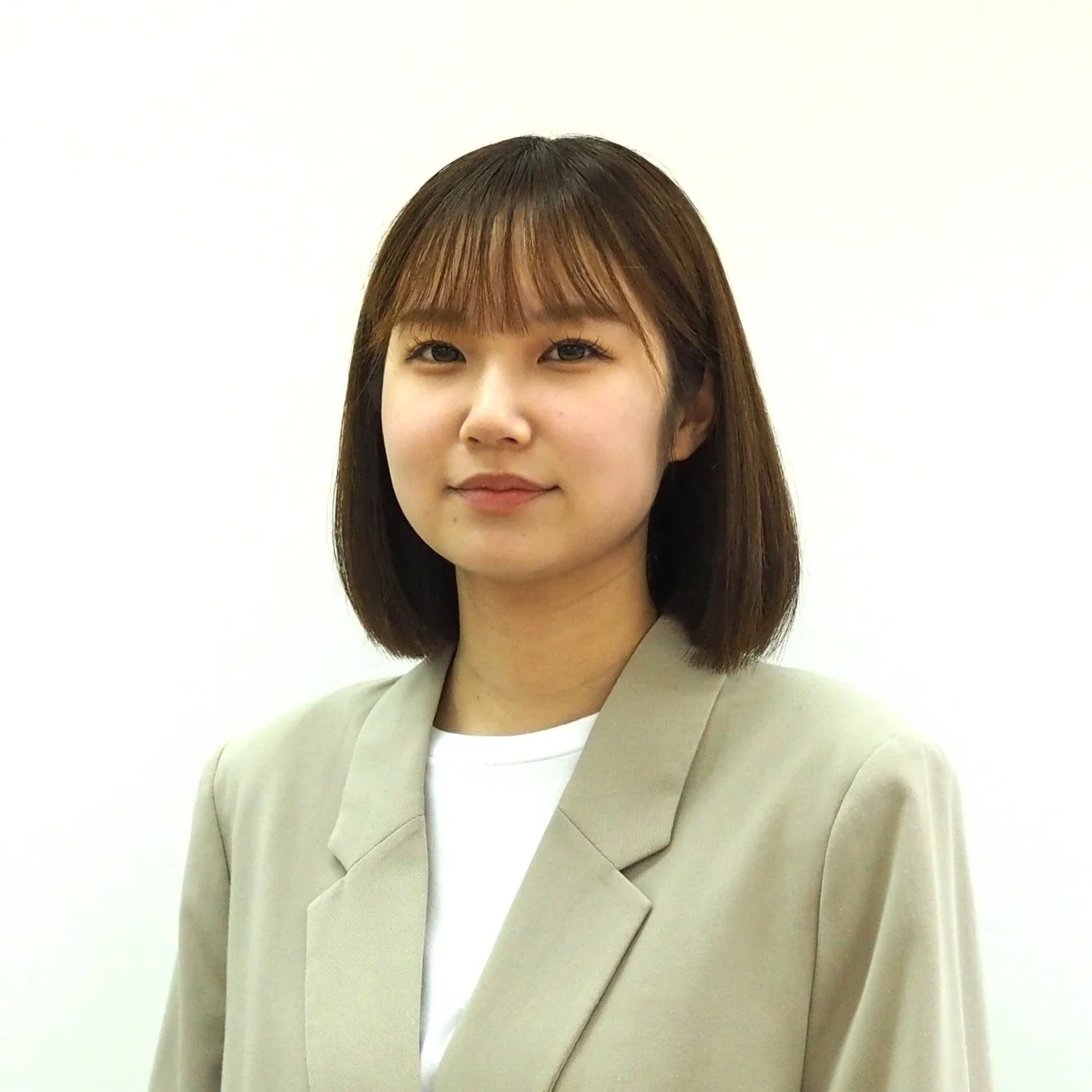 白い背景の前で、ベージュのブレザーと白いTシャツを着用したボブヘアの若い女性が正面を向いて立っています。穏やかで真面目な表情をしています。