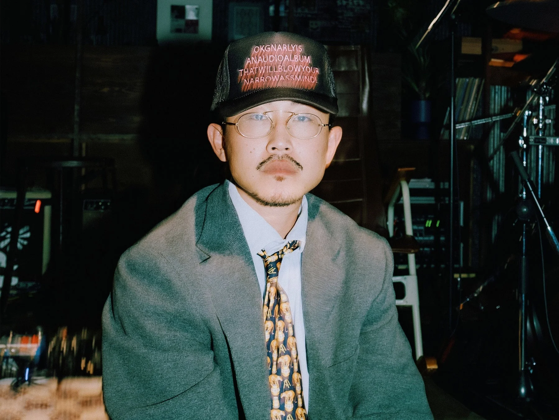 口ひげと顎ひげを生やし、眼鏡をかけた男性が、文字入りのキャップ、グレーのジャケット、柄物のネクタイを着用してカメラを見つめているポートレートです。背景は薄暗く、機材が置かれているようです。