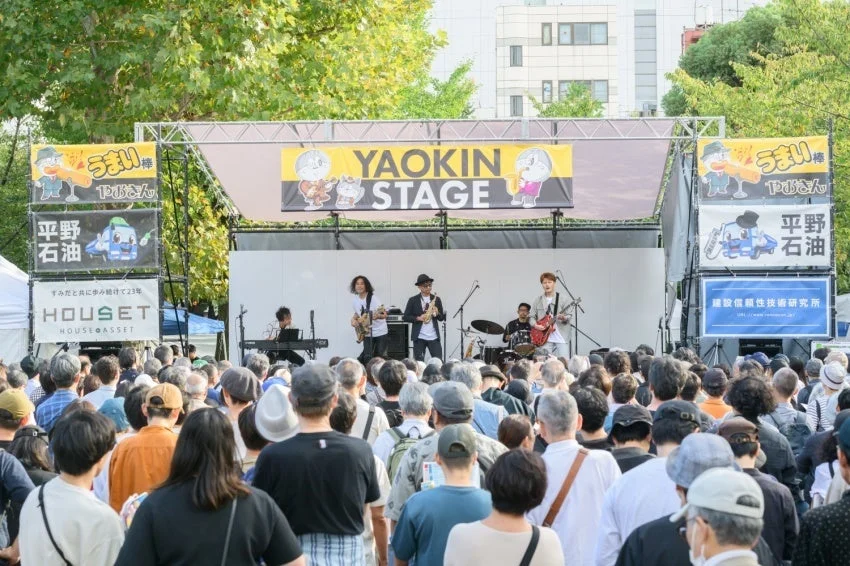 屋外ステージでバンドがライブ演奏を行い、大勢の観客が楽しんでいる風景です。ステージ上には「YAOKIN STAGE」の看板や協賛企業の広告が掲げられ、活気あるイベントの様子がうかがえます。