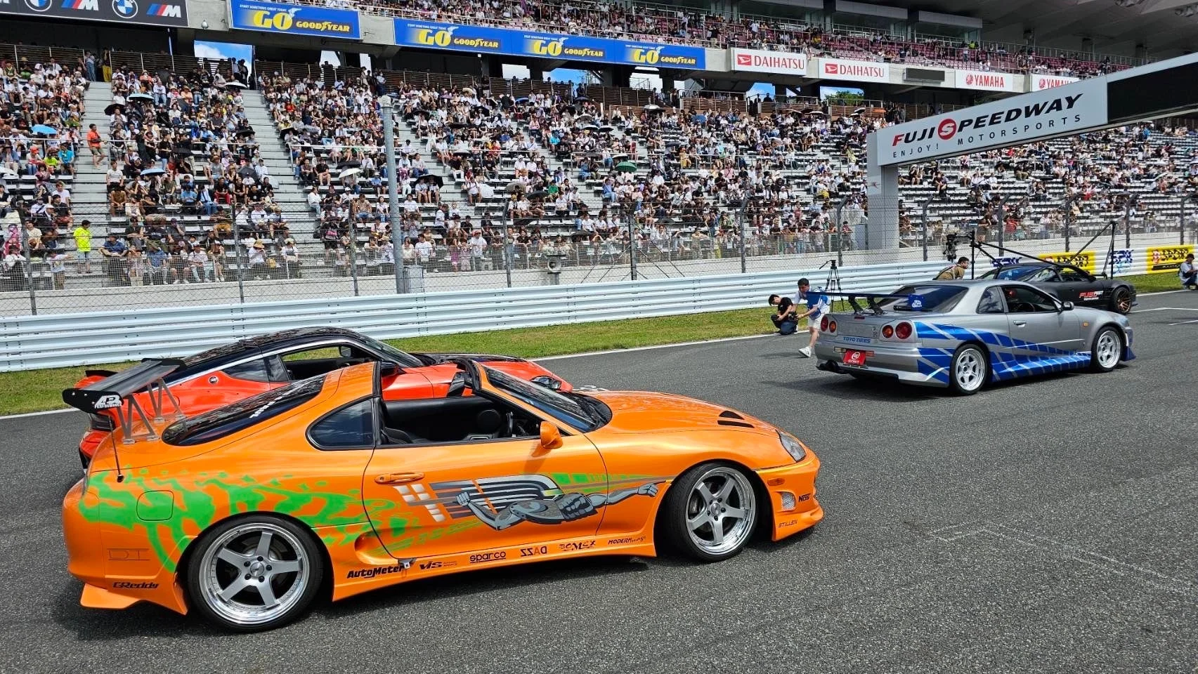 富士スピードウェイで開催されたモータースポーツイベントで、オレンジ色のトヨタスープラやシルバーの日産スカイラインGT-Rなど、カスタムされたスポーツカーが並ぶ様子が写っている。観客席には多くの人々が詰めかけている。
