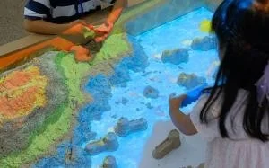 子供たちがインタラクティブな砂場で遊んでいる様子を捉えた画像です。一人の女の子が青く光る「水」の部分で遊び、もう一人の子供が色鮮やかな地形が投影された砂山を操作しています。光とプロジェクションマッピングを組み合わせた創造的な遊びの場面です。