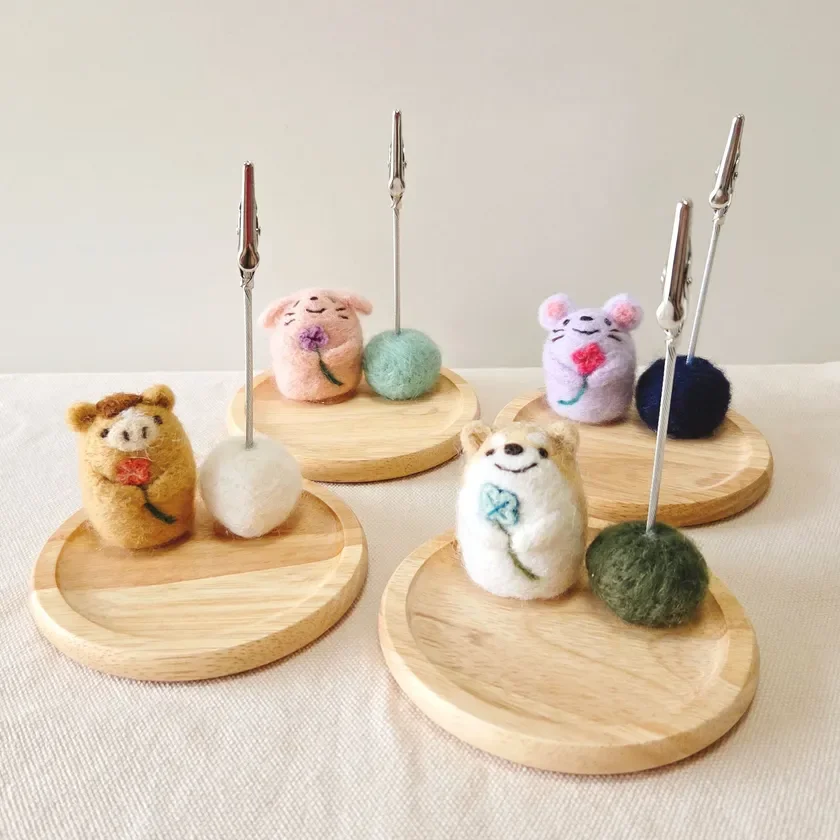 羊毛フェルトで作られた、ブタ、ウサギ、ネズミ、イヌなどの動物たちのメモスタンドが複数並べられています。それぞれの動物が花やクローバーを持っており、小さな木製の台座に固定されたクリップが特徴のかわいらしい手芸品です。