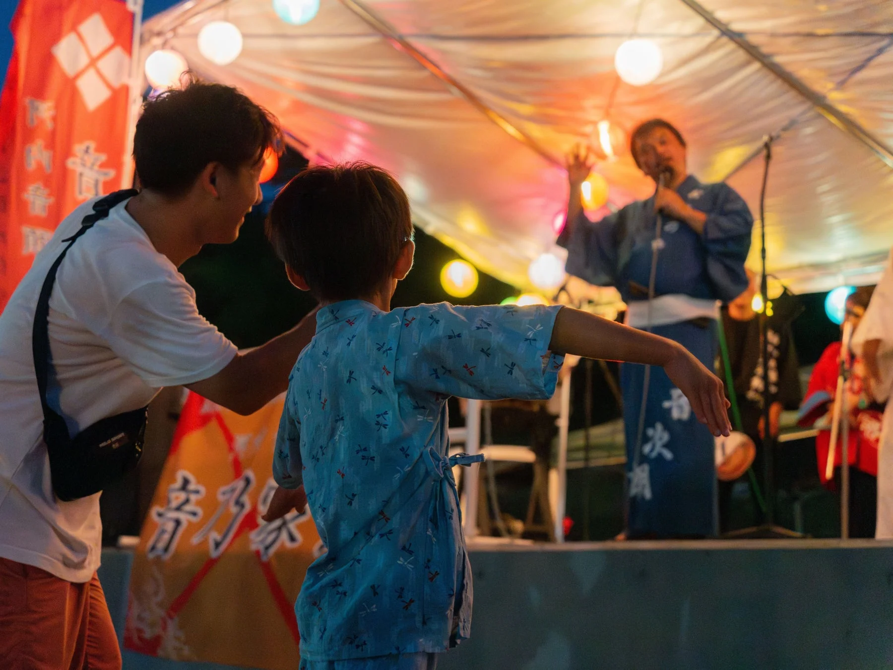 日本の夏祭り会場で、父親と子供が盆踊りを楽しんでいる様子です。子供は水色の浴衣を着て踊っており、ステージでは着物を着た大人がマイクを持って歌っています。会場は色とりどりの提灯で飾られ、賑やかな雰囲気が伝わります。