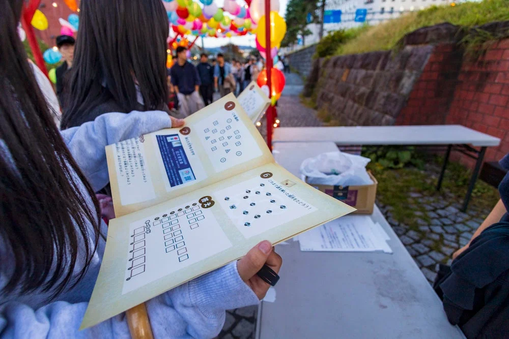 カラフルな提灯が飾られた屋外イベントで、一人の人物が世界遺産巡りのスタンプラリー冊子を広げています。謎解きのような要素もあり、参加者が楽しんでいる様子が伺えます。