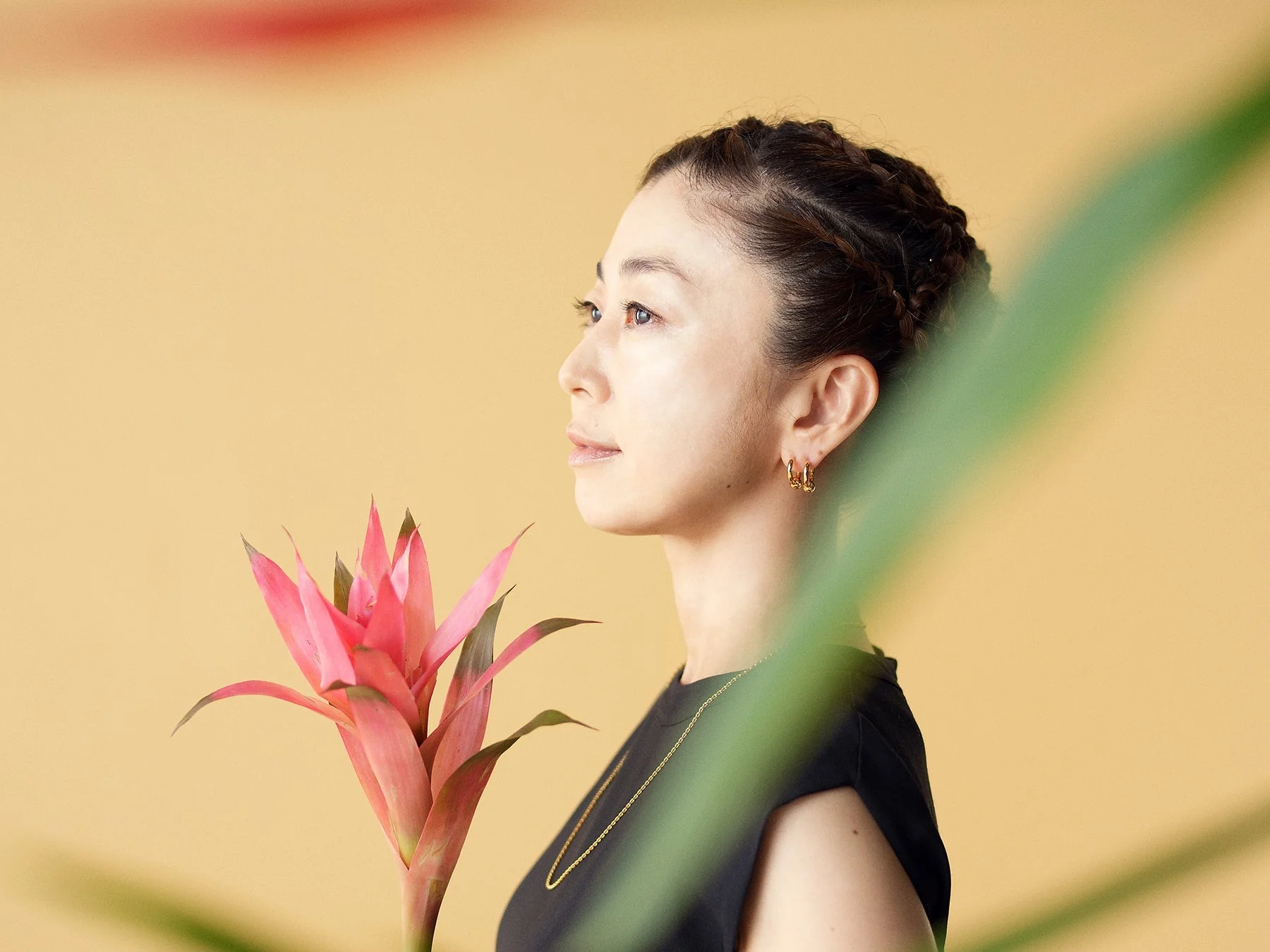 淡い黄色の背景を背に、横顔の女性が鮮やかなピンクのブロメリアの花と共に写っています。女性は金色のピアスとネックレスを身につけ、黒いトップスを着用。手前の緑の葉が奥行きを加え、自然で上品な雰囲気のポートレートです。