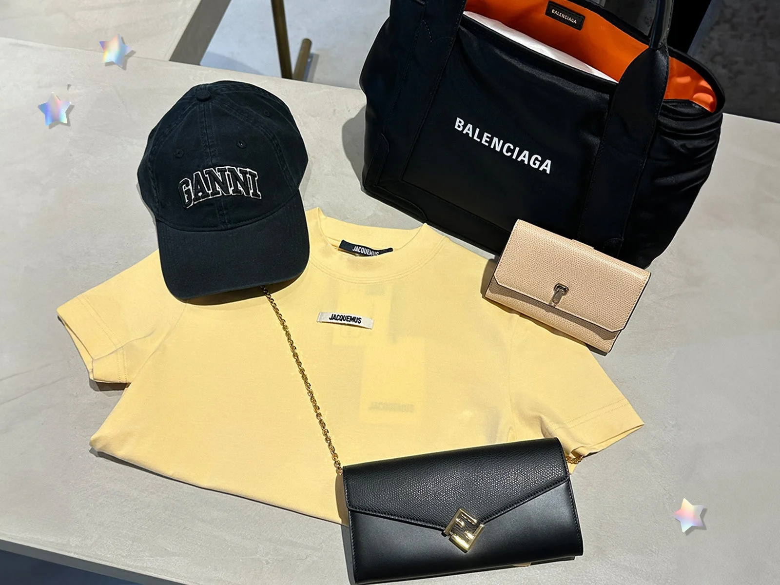 黒いキャップ、黄色いTシャツ、黒いトートバッグ、ベージュと黒の財布など、複数のブランド品が平置きされたファッションアイテムの集合写真