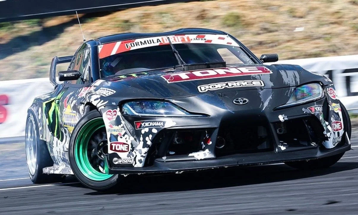 黒いボディに多数のスポンサーロゴが貼られたトヨタ・スープラのドリフトカーが、サーキットで白煙を上げながらドリフト走行している瞬間