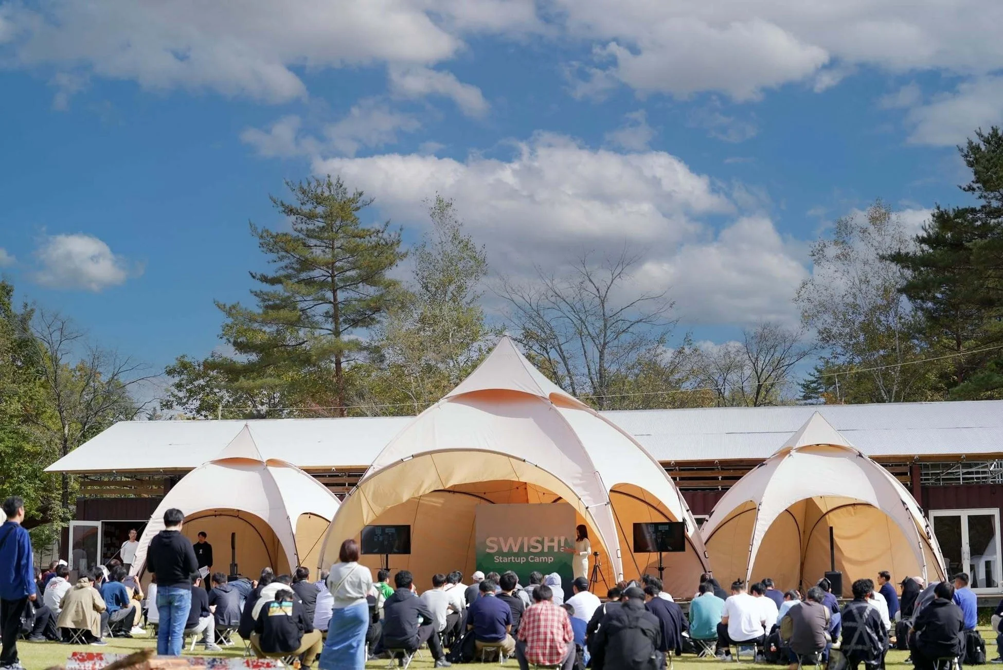 青空の下、屋外で開催されたスタートアップキャンプの様子。参加者が大きなテントの前に集まり、プレゼンテーションを熱心に聞いています。