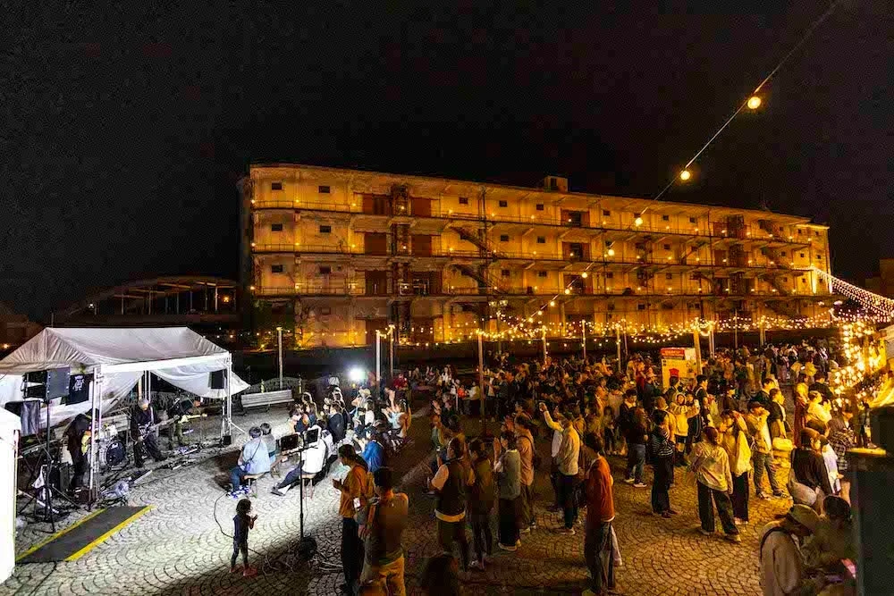 夜間に開催されている屋外イベントの様子です。ステージではバンドが演奏しており、多くの観客が音楽を楽しんでいます。背景にはライトアップされた大きな建物があり、会場全体が暖かな光で彩られています。