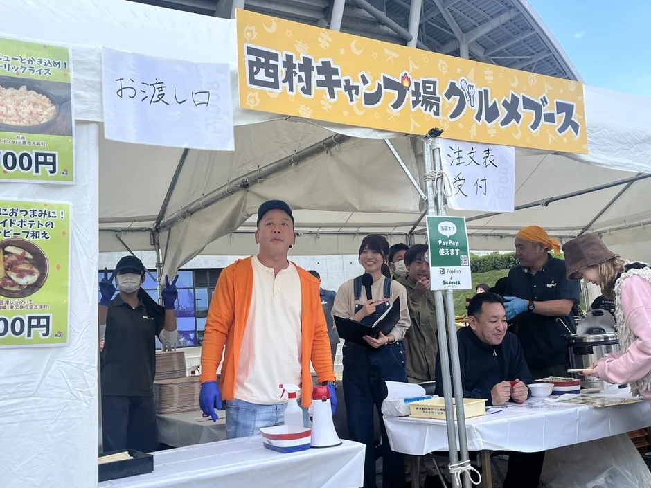 屋外イベント会場に設置された「西村キャンプ場グルメブース」の様子。オレンジ色のパーカーを着た男性を中心に、複数のスタッフが注文受付や料理の提供を行っています。PayPayの決済も利用可能で、多くの人々で賑わう活気ある飲食ブースの風景です。