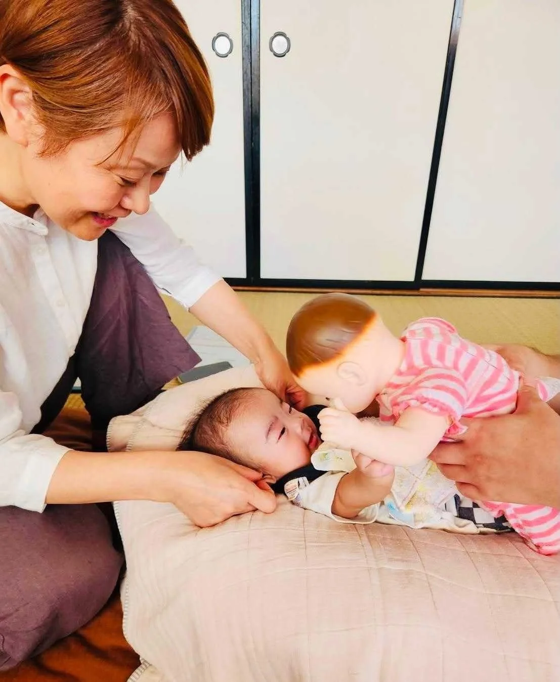 笑顔の女性が赤ちゃんを見守る中、別の大人が赤ちゃん人形を使って交流している様子
