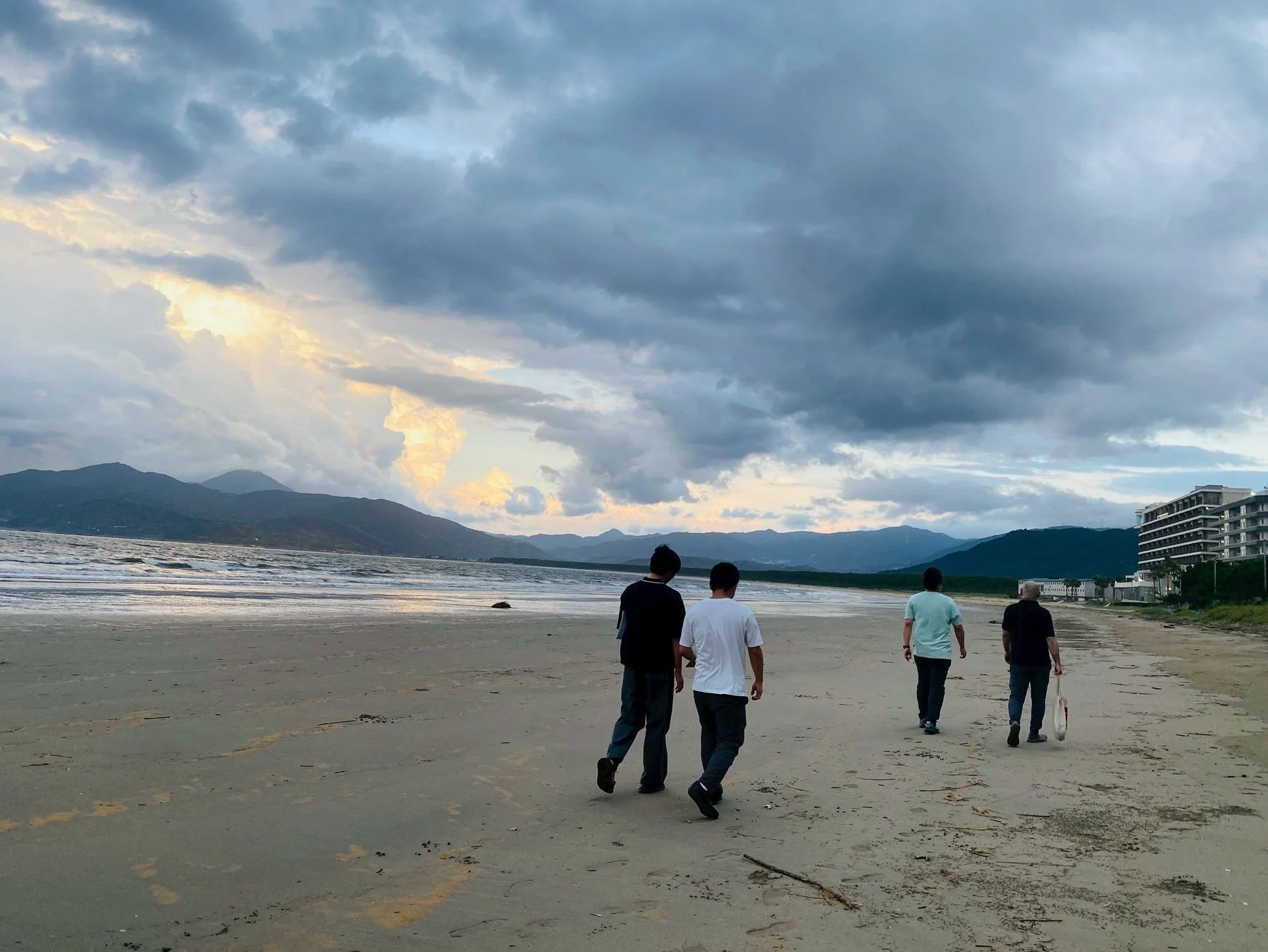夕暮れ時、波打ち際と遠くの山々を背景に、4人の男性が砂浜を歩く穏やかな風景