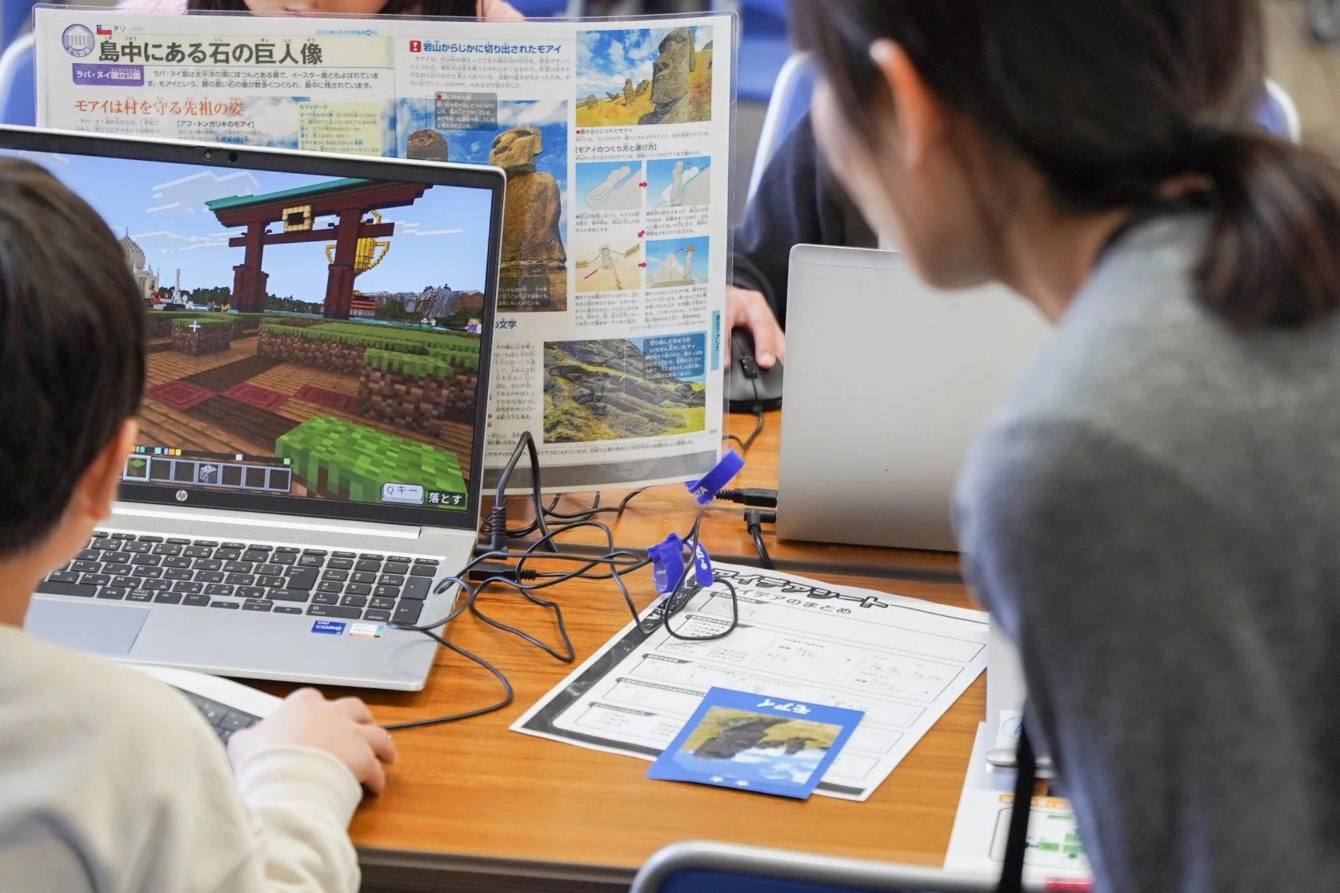 子供がノートパソコンでマインクラフトを操作し、モアイ像に関する学習資料が置かれている様子