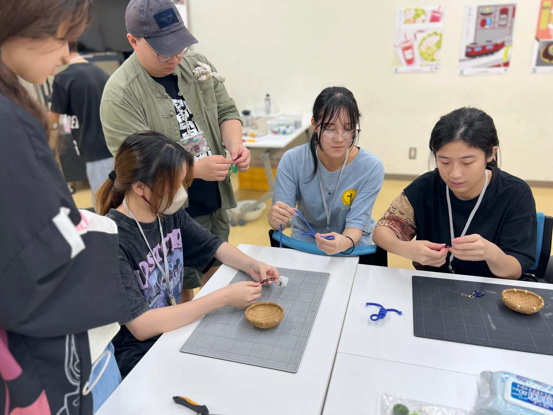 数人の若者が室内でクラフトワークショップに参加しており、ひもやビーズを使ってアクセサリーのような小物を制作している様子