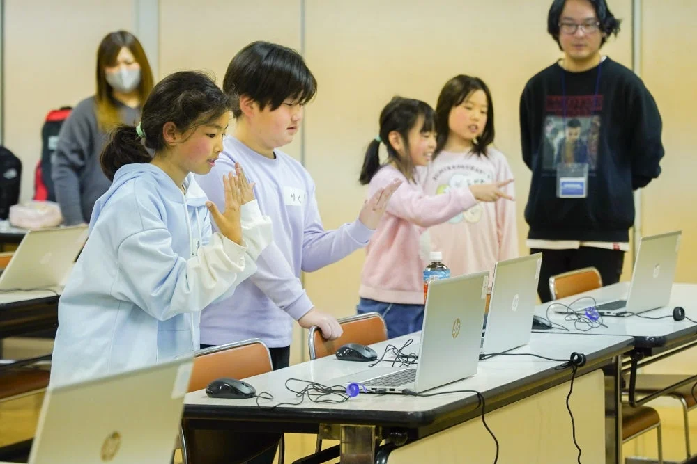 子供たちが複数台のノートパソコンを使って学習やプログラミングを行っているワークショップの様子